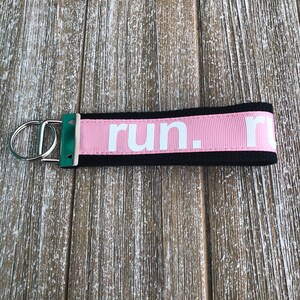 K&ouml;nnte beinhalten: Ein rosa-schwarzer Schl&uuml;sselanh&auml;nger mit dem Wort "run." in wei&szlig;en Buchstaben. Der Schl&uuml;sselanh&auml;nger hat einen silbernen Metallring und ein gr&uuml;nes Akzentst&uuml;ck. Der Hintergrund ist eine Holzoberfl&auml;che.