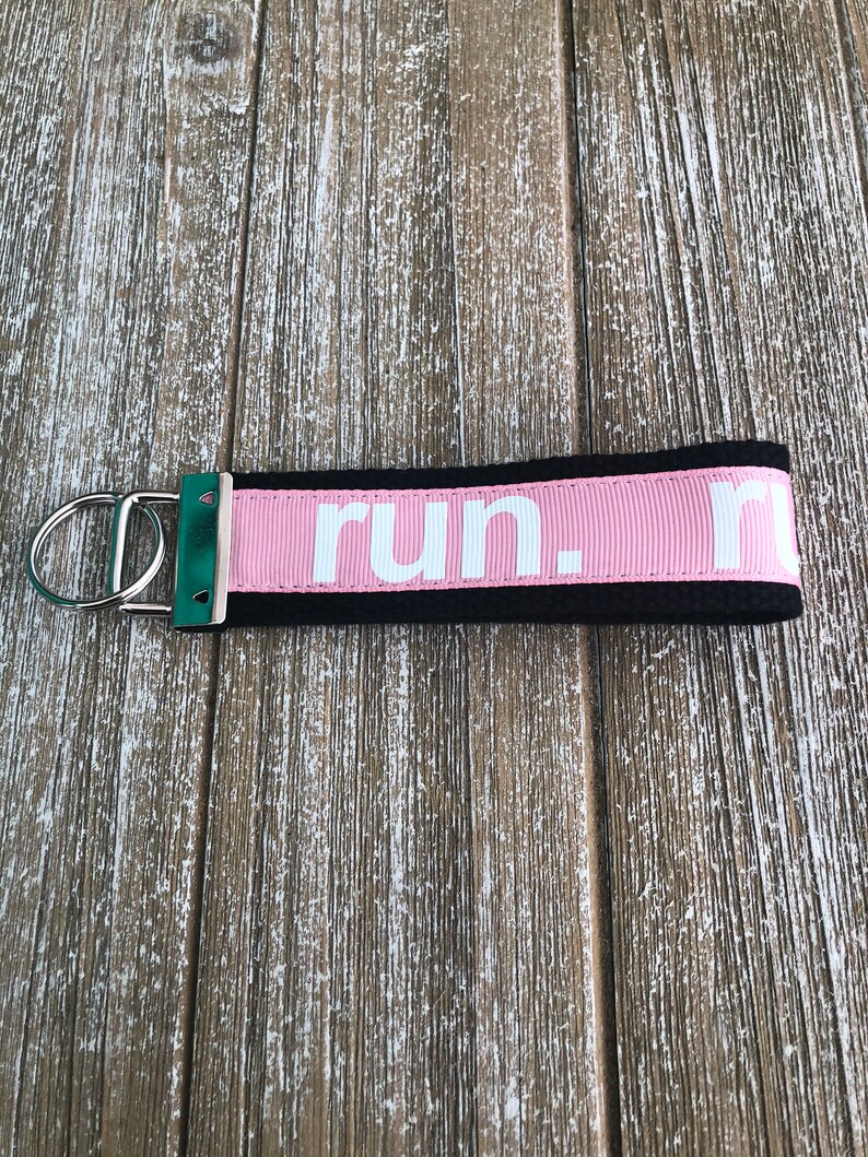 K&ouml;nnte beinhalten: Ein rosa-schwarzer Schl&uuml;sselanh&auml;nger mit dem Wort "run." in wei&szlig;en Buchstaben. Der Schl&uuml;sselanh&auml;nger hat einen silbernen Metallring und ein gr&uuml;nes Akzentst&uuml;ck. Der Hintergrund ist eine Holzoberfl&auml;che.