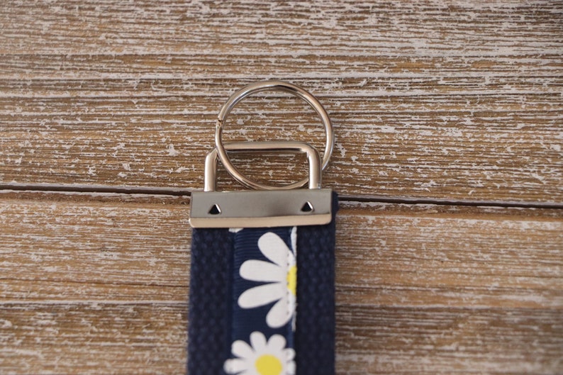 K&ouml;nnte beinhalten: Ein marineblauer Schl&uuml;sselanh&auml;nger mit einem G&auml;nsebl&uuml;mchenmuster. Der Schl&uuml;sselanh&auml;nger hat einen silbernen Metallring und ein Stoffband mit wei&szlig;en G&auml;nsebl&uuml;mchen und gelben Zentren. Der Hintergrund ist eine Holzoberfl&auml;che.
