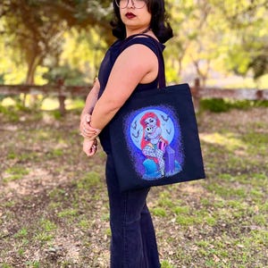 May include: Black tote bag featuring a cartoon illustration of a mermaid with red hair, sitting on a tombstone. The design includes a purple and blue color scheme with a full moon background. The bag is held by a person wearing black clothing.