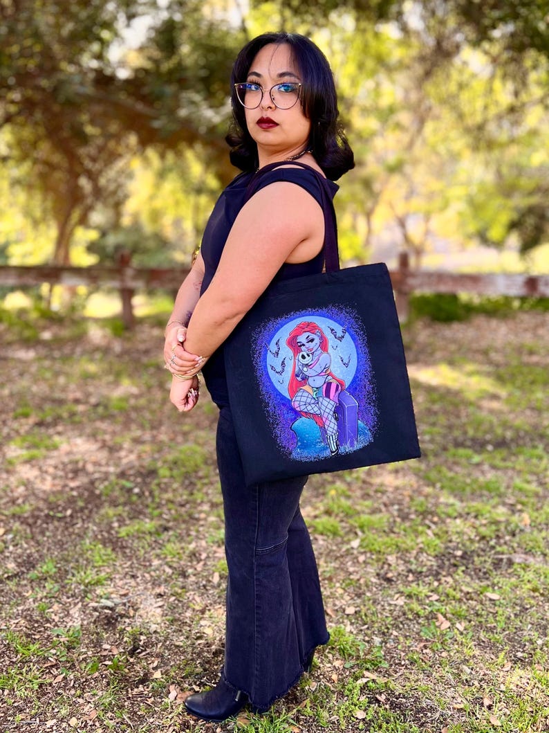 May include: Black tote bag featuring a cartoon illustration of a mermaid with red hair, sitting on a tombstone. The design includes a purple and blue color scheme with a full moon background. The bag is held by a person wearing black clothing.