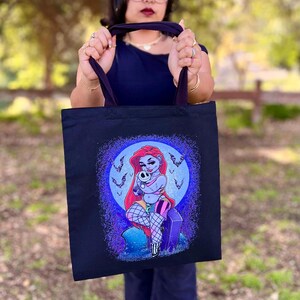 May include: Black tote bag featuring a colorful illustration of a cartoon character with red hair, holding a skull. The design includes a moon, bats, and a tombstone. The bag has dark purple handles.