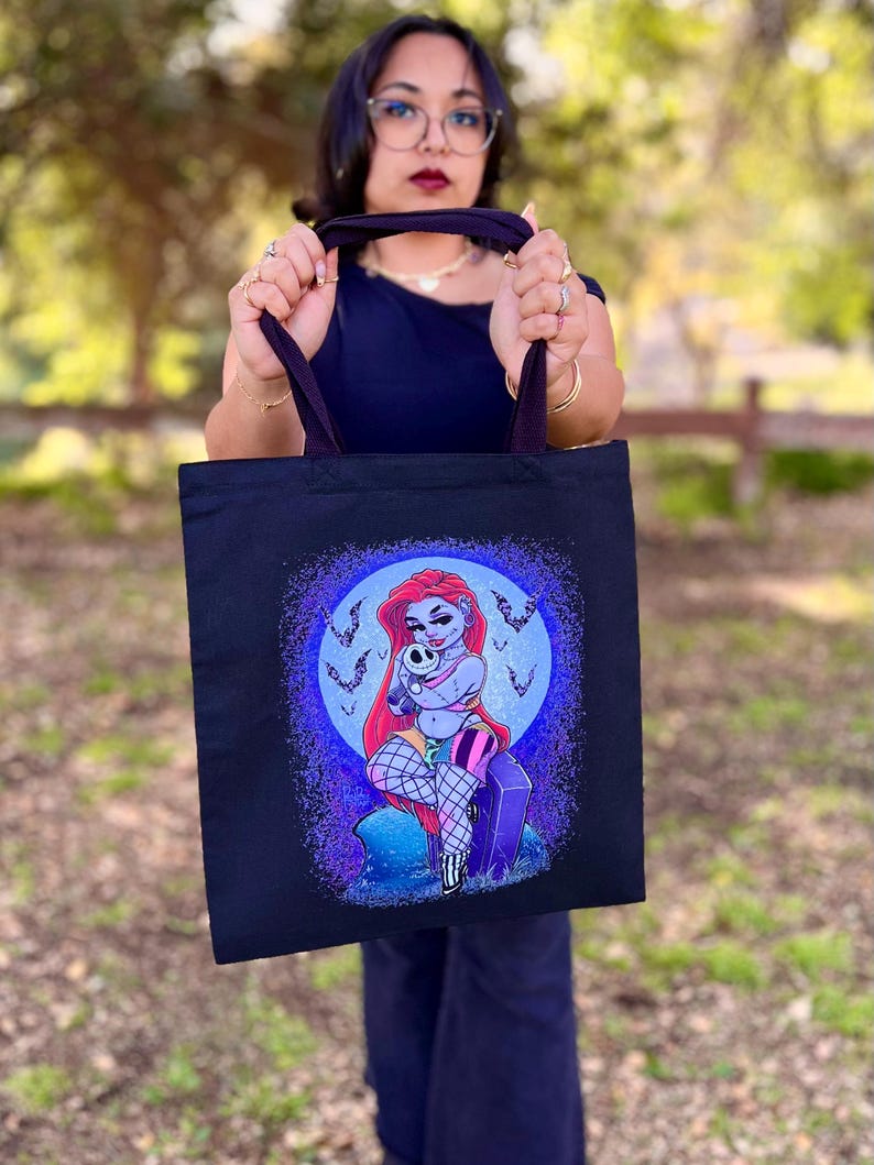 May include: Black tote bag featuring a colorful illustration of a cartoon character with red hair, holding a skull. The design includes a moon, bats, and a tombstone. The bag has dark purple handles.
