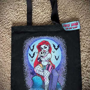 May include: Black tote bag featuring a colorful illustration of a woman with red hair, holding a skull, and sitting on a tombstone. The design includes a full moon, bats, and the text "TOTE BAGS BY REY MEZ" on a tag.