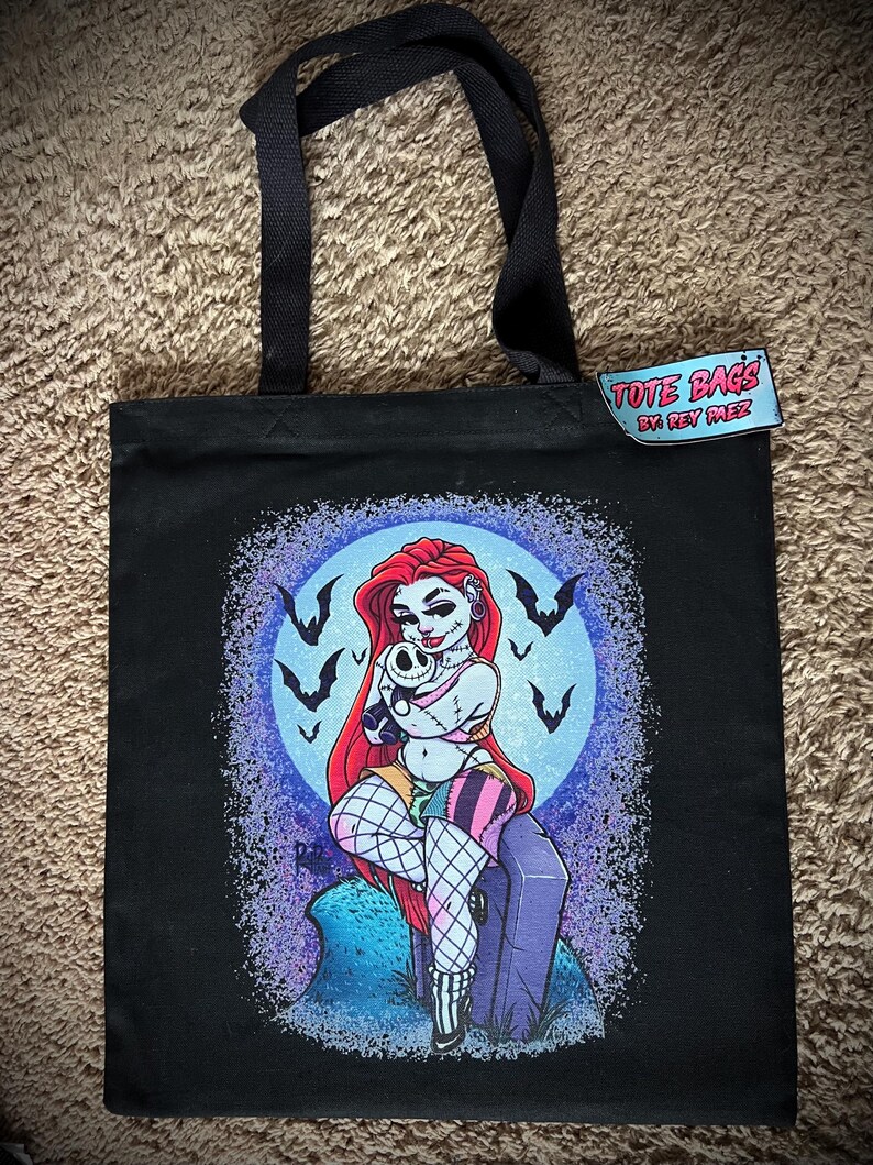 May include: Black tote bag featuring a colorful illustration of a woman with red hair, holding a skull, and sitting on a tombstone. The design includes a full moon, bats, and the text "TOTE BAGS BY REY MEZ" on a tag.
