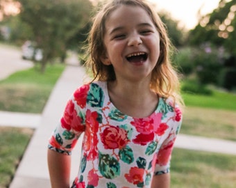 Peut inclure: Une jeune fille portant une robe blanche à imprimé floral et un motif géométrique sur la jupe. Elle sourit et a les cheveux tirés en arrière en un chignon.