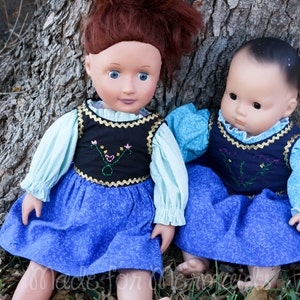 May include: Two dolls wearing blue and white dresses with embroidered vests. The dolls are sitting next to a tree trunk.
