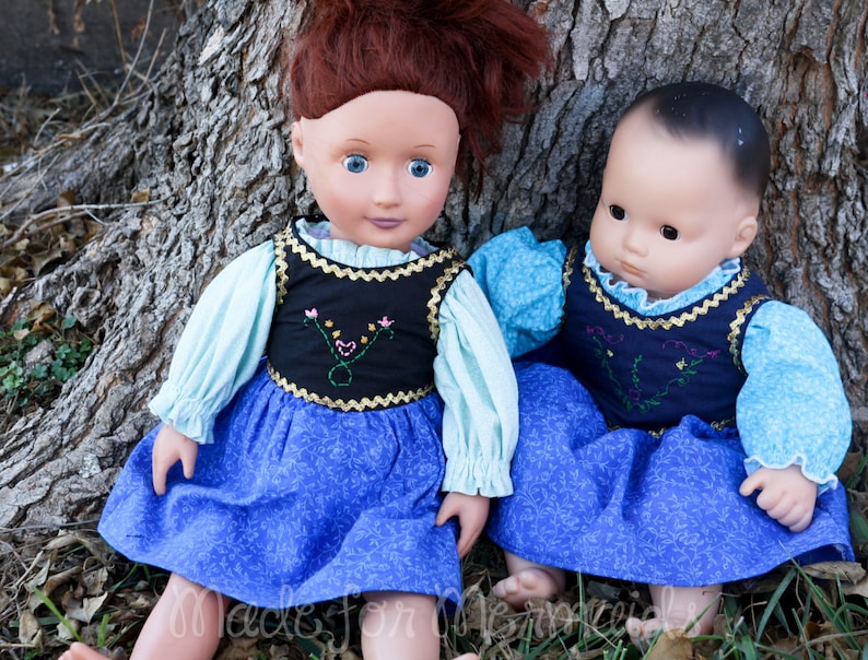 May include: Two dolls wearing blue and white dresses with embroidered vests. The dolls are sitting next to a tree trunk.