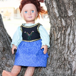 May include: A doll wearing a blue and black dress with a floral pattern. The doll has red hair and blue eyes. The doll is sitting against a tree trunk.