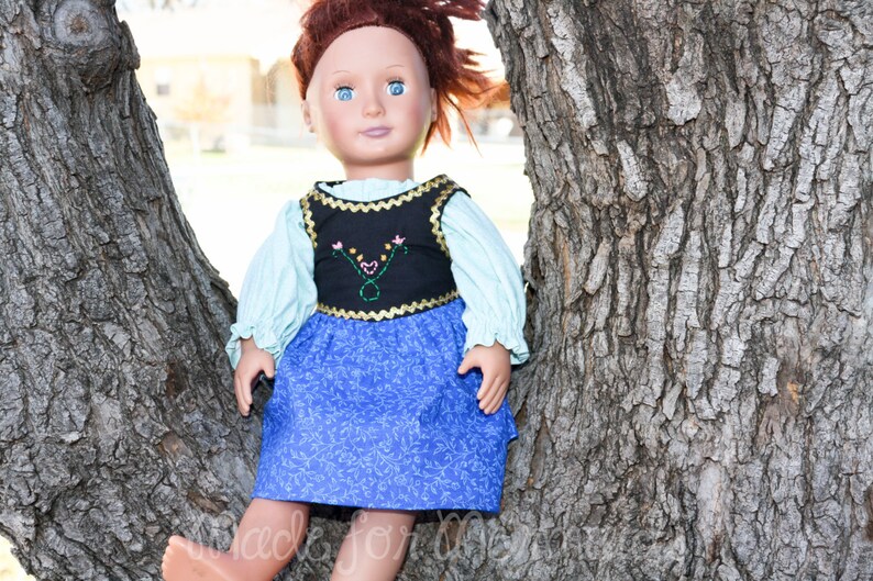 May include: A doll wearing a blue and black dress with a floral pattern. The doll has red hair and blue eyes. The doll is sitting against a tree trunk.