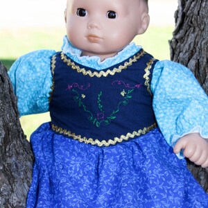 May include: A doll wearing a blue and white dress with a navy blue vest featuring embroidered floral details. The doll has a light brown complexion and dark brown hair.