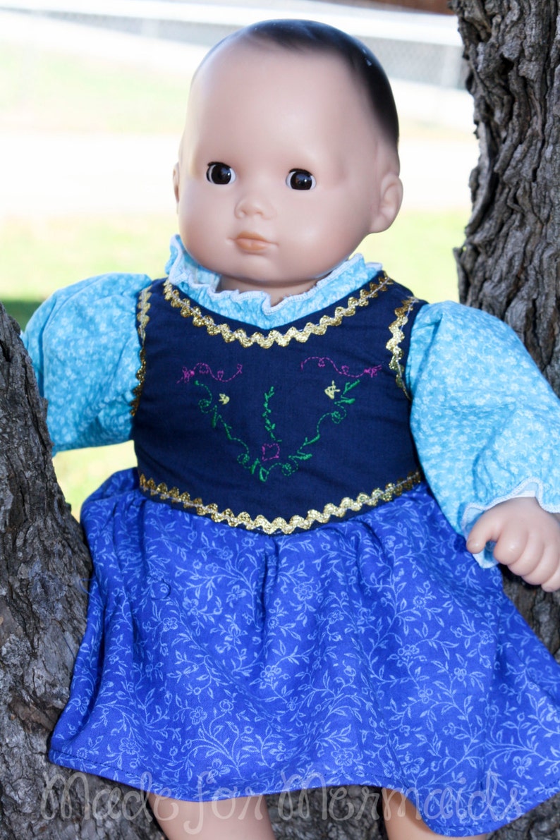 May include: A doll wearing a blue and white dress with a navy blue vest featuring embroidered floral details. The doll has a light brown complexion and dark brown hair.