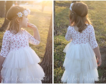 May include: A young girl wearing a white tulle dress with a floral print bodice and a flower crown. The dress has multiple layers of tulle and is a light, airy style. The girl is standing in a grassy field with trees in the background.