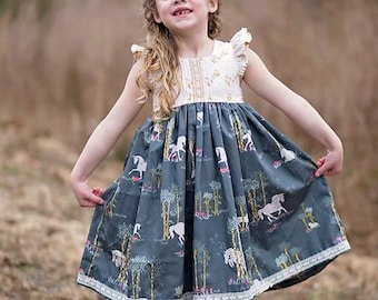 Peut inclure: Une jeune fille portant une robe blanche et bleue à imprimé licorne et une couronne de fleurs. Elle est debout dans un champ herbeux et sourit.