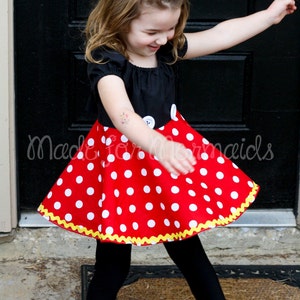 May include: A child's outfit featuring a black top with white buttons and a red polka dot skirt. The skirt has a yellow scalloped trim. The outfit is paired with black leggings and a red polka dot bow.