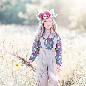May include: A young girl wearing a floral print blouse and beige overalls with a flower crown on her head. She is holding a bouquet of dried grass and standing in a field of tall grass.