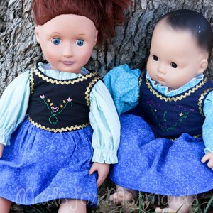 May include: Two dolls wearing blue dresses with floral patterns and white blouses. The dolls are sitting next to a tree trunk.