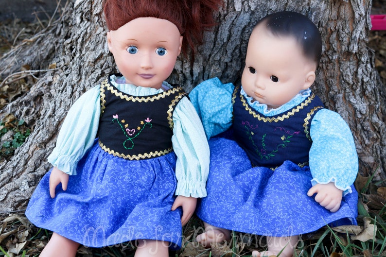 May include: Two dolls wearing blue dresses with floral patterns and white blouses. The dolls are sitting next to a tree trunk.