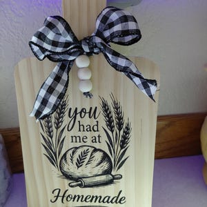 May include: A wooden cutting board with a handle, decorated with a black and white checkered bow and three white beads. The board features a graphic of a loaf of bread, wheat stalks, and the text "you had me at Homemade."