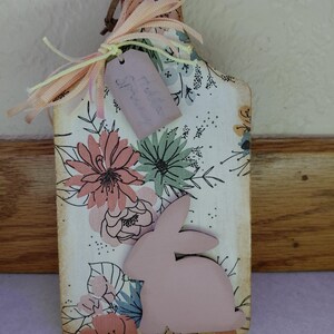 May include: A decorative wooden cutting board with a floral design and a pink bunny silhouette. The board has a handle with a ribbon and a tag that says "Spring". The color scheme includes pink, green, and blue.