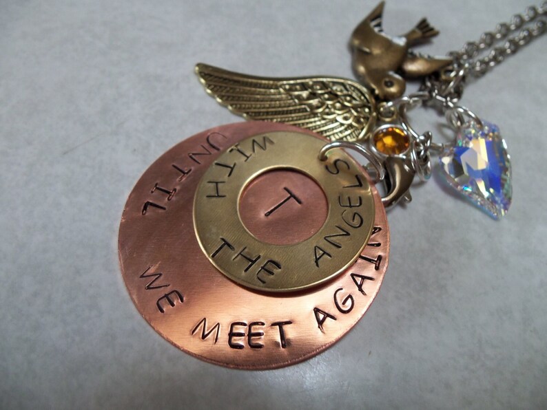 May include: A copper and brass necklace with a charm that reads "We Meet Again With The Angels". The charm is attached to a silver chain with a small bird and crystal charm.