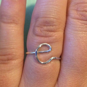 Puede incluir: Un anillo plateado con la forma de la letra "e" se muestra en un dedo. El anillo es delicado y parece estar hecho de alambre. El fondo es de color verde claro.