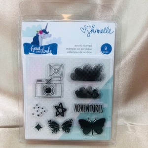 May include: A clear plastic package containing 9 acrylic stamps with a unicorn illustration and the text "Head in the Clouds". The stamps include a camera, clouds, stars, a starburst, and butterflies. The package also includes the text "Shimelle" and "Adventures".