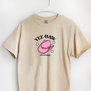 May include: A beige t-shirt with a pink cowboy hat graphic and the text "Yee Haw" and "chi omega" printed on it.