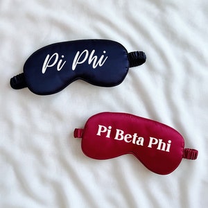 Puede incluir: Dos antifaces de satén, uno negro con texto blanco "Pi Phi" y otro burdeos con texto blanco "Pi Beta Phi".
