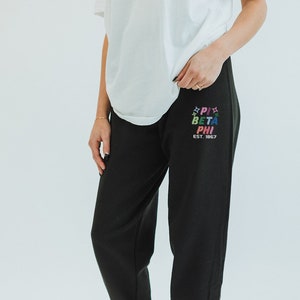 Könnte beinhalten: Schwarze Jogginghose mit einem bunten "Pi Beta Phi Est. 1867" Aufdruck auf dem linken Bein.