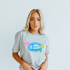 May include: A grey t-shirt with a blue oval graphic that says "CHI OMEGA" with a yellow flower, pink smiley face, and green lightning bolt.