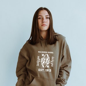 May include: A brown hoodie with a white graphic of a playing card queen with the text "Phi Sigma Sigma" and "Lucky You" on either side of the card. The text "Est. 1913" is below the card.