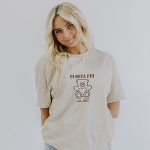Könnte beinhalten: Ein hellbeiges T-Shirt mit einem braunen Teddybär-Motiv und dem Text "Pi Beta Phi est. 1867".