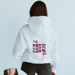Könnte beinhalten: Weißer Kapuzenpullover mit einem pinkfarbenen und violetten Aufdruck, der dreimal "Pi Beta Phi" lautet. Der Aufdruck enthält auch rosa Sterne.