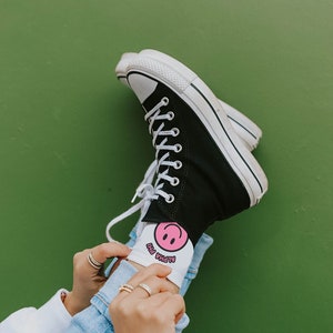 以下が含まれることがあります： 白の紐と白のソールが付いた黒いハイカットスニーカー。人は、ピンクのスマイリーと「ALPHA PHI」の文字が書かれた白い靴下を履いています。