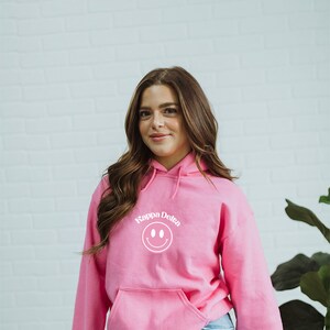 Puede incluir: Una sudadera con capucha rosa con una carita sonriente blanca y el texto "Kappa Delta" en un arco blanco sobre la carita sonriente.