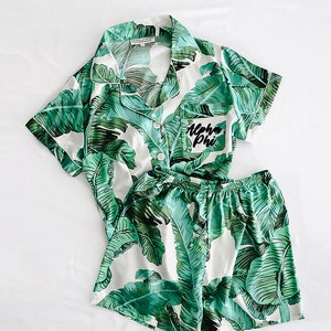 Può includere: Pigiama bianco con stampa a foglie tropicali verdi. La parte superiore ha un colletto abbottonato e maniche corte. I pantaloncini hanno una cintura elastica. La tasca sulla parte superiore ha il testo "Alpha Phi" ricamato.