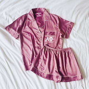 May include: A pink satin pajama set with short sleeves and shorts. The top has a white pocket with the letters "AΦ" embroidered on it. The label on the top reads "Spikes and Seams".