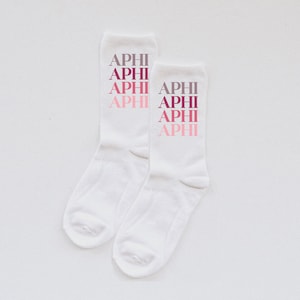Puede incluir: Dos pares de calcetines blancos con la palabra "APHI" repetida cuatro veces en cada calcetín en un degradado de rosa, rojo y gris.