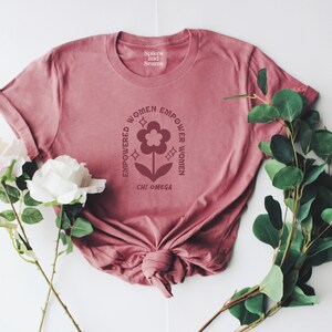 May include: A pink t-shirt featuring a graphic design of a flower with the text "Empowered Women Empower Women" and "Chi Omega" printed on it.