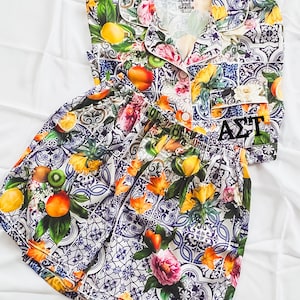 May include: A white and blue patterned pajama set with a short-sleeved top and shorts. The top has a button-up closure and a collar. The shorts have an elastic waistband. The pattern features fruit, flowers, and geometric designs. The top has a label that reads "Splkes and Seams".