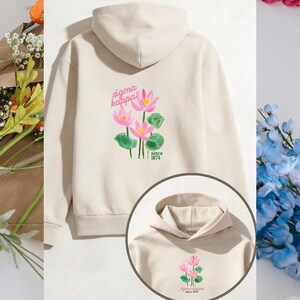 Puede incluir: Una sudadera con capucha beige con un diseño floral de flores rosas y hojas verdes. El texto "sigma kappa" y "since 1874" están impresos encima y debajo de las flores. La sudadera tiene capucha y mangas largas.