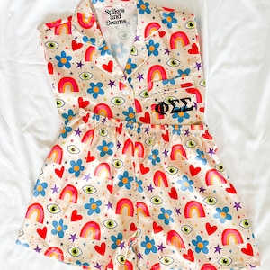 Puede incluir: Un conjunto de pijama de color crema con una parte superior sin mangas y pantalones cortos. La tela presenta un estampado colorido de arcoíris, estrellas, corazones, flores y ojos. La parte superior tiene un cuello abotonado y un bolsillo con las letras "ΦΣΣ" en él. La etiqueta en la parte superior dice "Spikes and Seams".