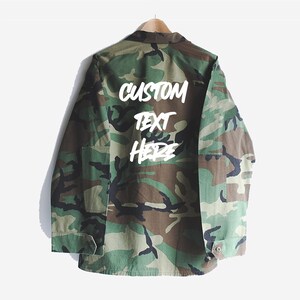 Puede incluir: Una chaqueta verde de camuflaje con las palabras "CUSTOM TEXT HERE" escritas en blanco en la espalda.