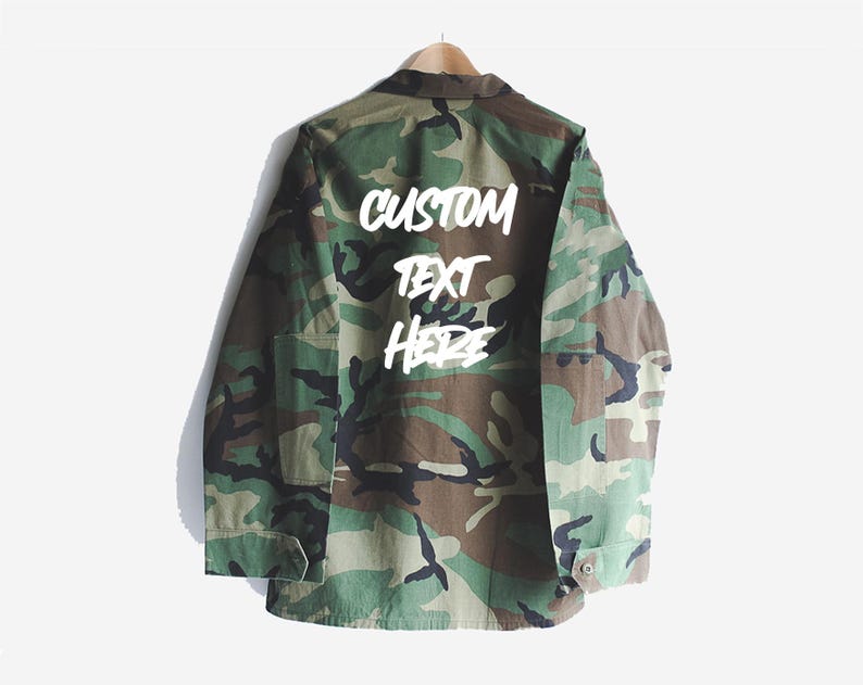 Puede incluir: Una chaqueta verde de camuflaje con las palabras "CUSTOM TEXT HERE" escritas en blanco en la espalda.