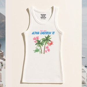 Puede incluir: Camiseta sin mangas blanca acanalada con un diseño tropical de palmeras, flores rosas, una tortuga y un cangrejo. El texto "ALPHA OMICRON PI" está en azul, con "EST. 1897" en rosa. La marca "Spikes and Seams" está impresa en la parte superior.