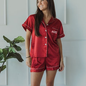 May include: A red satin pajama set with short sleeves and shorts. The top has a white button closure and a white embroidered "AOPI" on the left chest.