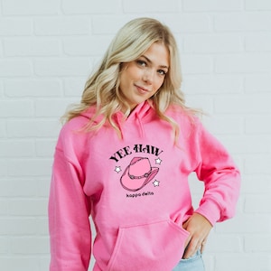 Puede incluir: Sudadera rosa con bolsillo de canguro, con un diseño de sombrero de vaquero y el texto "Yee Haw Kappa Delta" en negro, con estrellas. La sudadera es de color rosa liso.