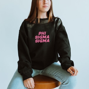 May include: A black sweatshirt with a pink "PHI SIGMA SIGMA" graphic print. The sweatshirt is being worn by a person sitting on a brown stool.