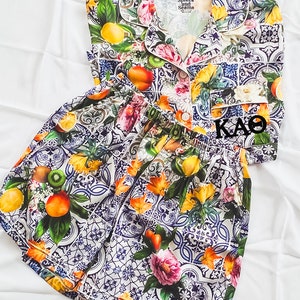 Peut inclure: Un ensemble pyjama blanc et bleu à motifs avec un haut à manches courtes et un short. Le haut a un col et une fermeture à boutons. Le motif présente une variété de fruits, de fleurs et de carreaux. Le texte "Spikes and Seams" est imprimé sur le haut.